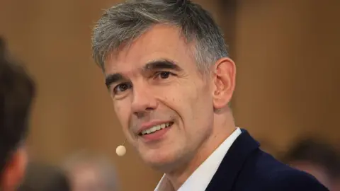 Matt Brittin wearing a suit on stage at a conference wearing a small microphone next to his cheek