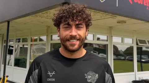BBC/Jasmine Lowe A tall man with bushy and curly hair, he is smiling at the camera and looks tired after a training session. He is also squinting due to the sun being in his eyes. He's wearing a black Hull City training top and standing in front of a single-story building with banks of glass windows.