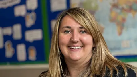 A woman is smiling at the camera. She has long blonde hair and is wearing a dark jacket. She's sitting in a classroom and behind her is a map of the world on the wall.