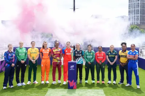 PA The 12 captains of the 2026 Women's T20 World Cup stand in a line on artificial grass behind the tournament trophy, each wearing their national kit, with pink and white smoke rising behind them.
