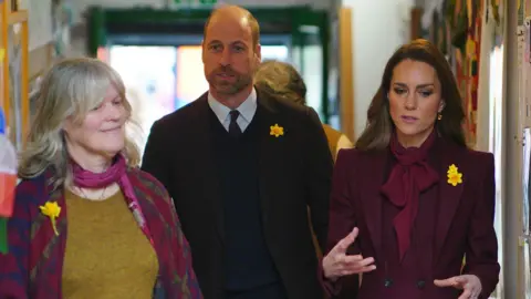 Reuters William and Catherine walking into the centre alongside a female member of the public who is guiding them in. She wears a green jumper and pink and purple scarf. William is in the middle in a shirt and tie and blazer and Catherine walks next to him, hands gesticulating as she talks