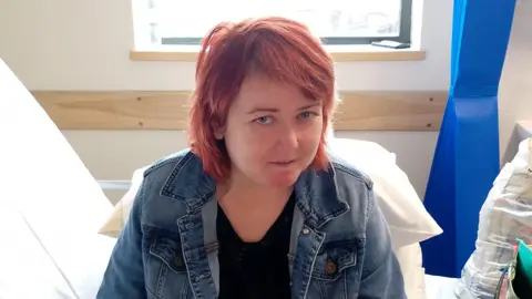 Family handout Alicia looking at the camera. She has short red hair and is wearing a black top under a blue denim jacket. She is sitting on a bed in a hospital or clinic's room, two large white pillows are sitting behind her. Further back is a window.