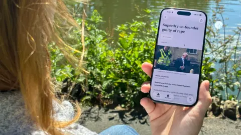 A woman looks at a BBC news story on a mobile phone. You can only see her hair and hand. The headline reads: Superdry co-founder guilty of rape.