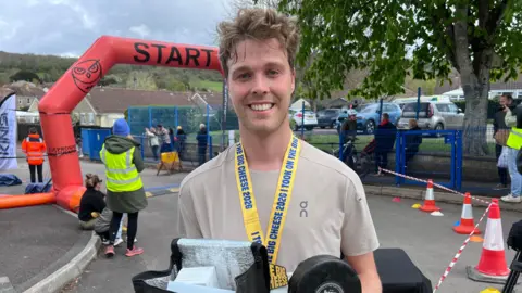 Sam Rodda smiles at the camera as he holds a hamper of cheese in his hands. He is wearing a beige running top with a yellow medal around his neck. He has short curly brown hair.