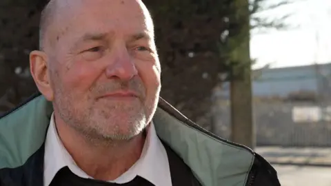 Tim Foster is seen standing outdoors, wearing a white shirt and dark jumper under a jacket, with a metal fence and trees behind him.
