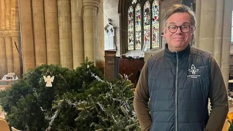 Jess Lord / BBC Father Stuart Cradduck, Rector at St Wulfram's in Grantham stands in front of the fallen Christmas tree he is looking at the camera and wearing, glasses, a blue gilet and a brown shirt