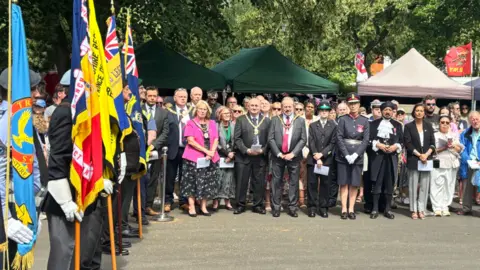 People are holding flags of various colours on the far left of the photo. In the centre and on the right, dozens of people are facing the camera. 