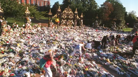 Getty Images A sea of flowers on the gates, railings and ground outside the Kensington Palace gates. A number of people are in the foreground of the picture laying more bouquets.  Kensington Palace can be seen through trees in the background beyond the gates.
