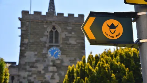 A church can be seen slightly blurred in the background with a blue clock on it and conifers in front of it. A sign can be seen close to the camera with a green and yellow arrow and a yellow circle which says Viking Way with a Viking helmet symbol on it.