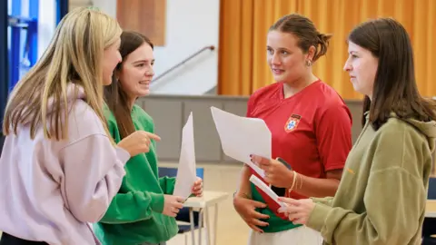 Four young women smile as they compare A-level results. They wear t-shirts and sweatshirts and are standing in their school hall. 