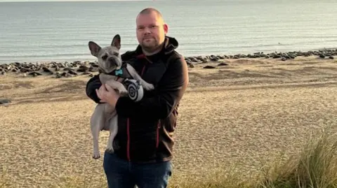 A man dressed in a black hoodie stood on a beach holding his dog