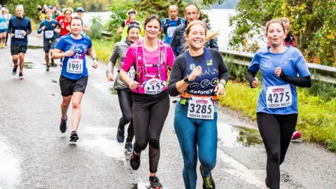 Runners smile for the photographer as they run down a rain-soaked road on the shores of Loch Ness. They have their entry numbers pinned to the front of their tops.