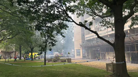 Smoke can be seen in the area with trees surrounding a concrete building which is cordoned off by police tape. 