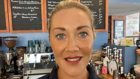 Brooke has dark blonde hair pulled back from her face, she has light blue eyes and behind her is her cafe's barista machine along with mugs and syrups. On the blue wall behind her is a number of black chalkboards listing what is sold there, along with prices.