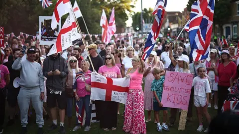 Henry Nicholls/AFP via Getty Images Large crowd of people waving Union Jack and St George flags outside council offices in Epping on 8 August 2025