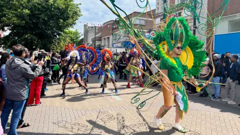 A man dressed in an enormous green feathered costume dances ahead of a group of women in red and blue outfits as people lining town centre streets watch them