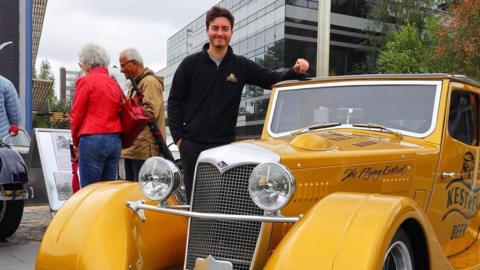Joel McNally with short dark hair standing next to a yellow car