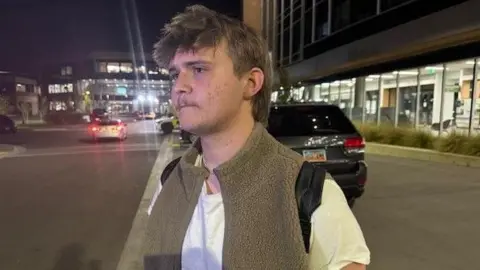 Brock Anderson, whose hair is in a mullet style, speaks to the BBC outside the university campus at night