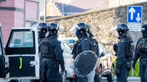 A number of police officers in black uniform and wearing helmets hold shields as they stand in a group. The open door of a police vehicle is to the left of the image