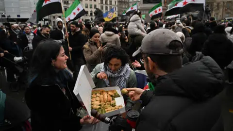 Getty Images An image of the demonstration at Trafalgar Square where people hold Syrian flags in the background. A woman hands out baklava and another woman and man reach in to take a bite.