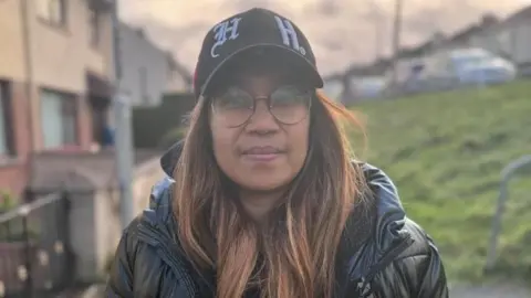 Cadalia Freitas - a woman with long, highlighted hair is wearing a baseball cap and circular glasses. She is also wearing a puffed black coat. She is standing on a footpath in front of a row of terraced houses.