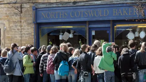 Dozens of people, several with rucksacks on, can be seen crowding around the entrance to a bakery in Bourton on the Water. The blue shop front reads "Norths Cotswolds Bakery".