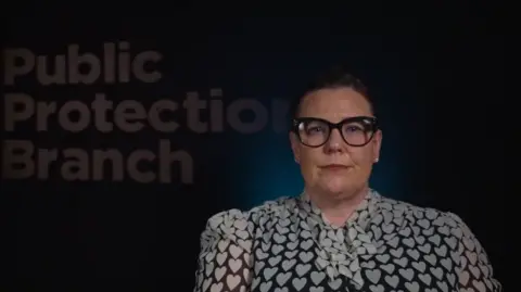 PSNI Det Ch Supt Zoë McKee is looking at the camera. She wears glasses. She's wearing small pearl earring. In the background, a board states public protection branch.