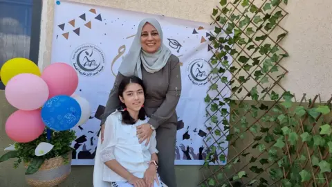 Shatha Abusalim Smiling mother Shatha stands over her seated daughter, Lamis, who is also smiling, at a school celebration in 2023. The picture is an exterior shot and the pair are infront of a banner with the school's name. There is a cluster of colourful balloons and green foliage. 