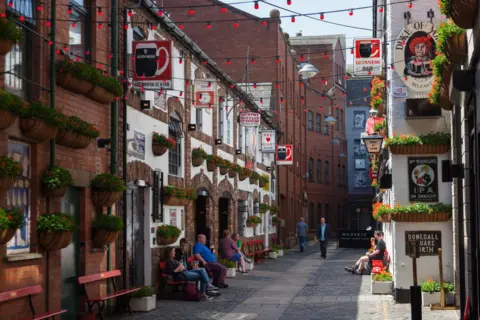 Getty Images Exterior of a red brick and white bar with numerous vintage alcohol adverts and hanging plants, there are people sat on red benches outside the venue, which is situated on a narrow cobbled street with red festoon lighting linking the buildings.