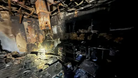 A hospital room that has been blackened and destroyed by fire. The ceiling appears to have collapsed and debris is strewn around the room. 
