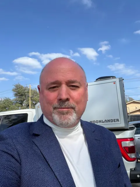Courtesy of John Erik Ege A man stands in front of a white pickup truck. He is wearing a turtleneck and a blue blazer and has a beard. 