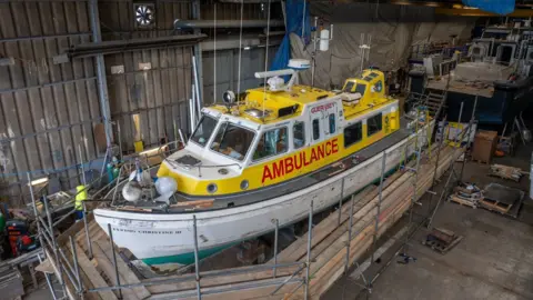 The vessel is bright yellow with a white hull. It has AMBULANCE written across the side in red capital letters. The boat is positioned in an industrial unit with scaffolding surrounding its hull. 