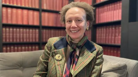 A woman in a green, navy and red checked jacket, with a similarly patterned scarf smiles at the camera. She has short, brown and curly hair and is sat on a grey sofa in front of bookcases filled with red, leather bound books.