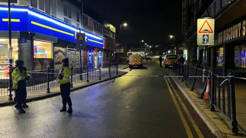 Nathan Turvey/BBC Two police officers stand in front of blue and white police tape in Dewsbury town centre.