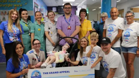 Wallace and Gromit's Grand Appeal A large group of medical staff in a hospital children's ward centred around a young child in a stroller and Teddy with his parents wearing t-shirts with logos for the Great Bristol Mini Run on them. 