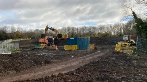 An orange JCB and two other yellow digger trucks working on a muddy piece of land. There are blue shipping containers on the side and yellow barrier fencing. 