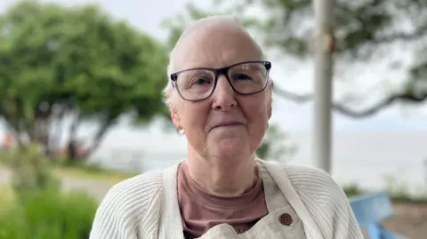 Sally is looking directly at the camera with a neutral expression on her face. She is wearing a cream cardigan, brown t-shirt and light brown dungarees. She has short white hair and glasses.