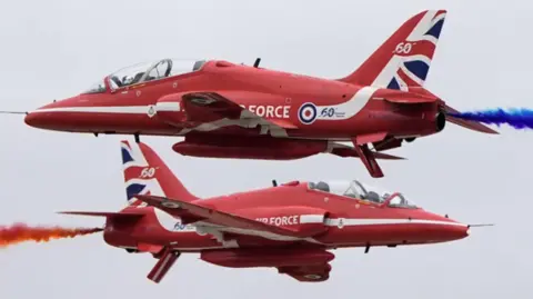 Two Red Arrows jets, travelling in opposite directions, pass each other within a whisker. One aircraft trails blue smoke, the other red smoke.