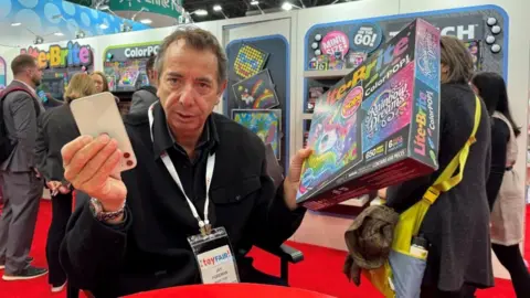 Jay Foreman holds up a mobile phone and a Lite-Brite board game at the North American Toy Fair