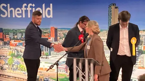BBC/Simon Thake A man with a closed shaved beard and brown hair is wearing a dark suit. He is shaking hands with a woman with blond hair, glasses and a brown suit on a podium. In the background are two men in dark suits with red and yellow rosettes on.