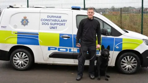 Dan Cuthbertson A black police dog with his handler stood in front of a police van