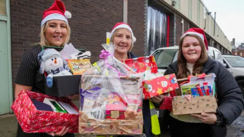 Three women are holding Christmas hampers. The boxes are wrapped in festive wrapping paper and include sweets, handwritten letters and teddies. The women are wearing red and white Santa hats.