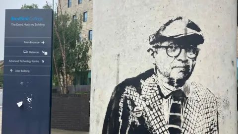 A graffiti image of David Hockney on a pillar with a sign for Bradford College behind