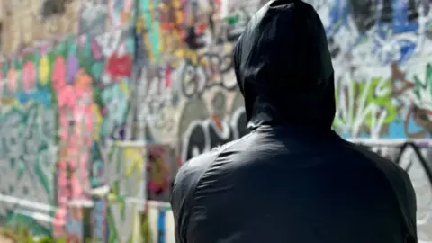 A mid shot of a person wearing a jacket with their hood up. They are standing in front of a wall which has been covered with graffiti.