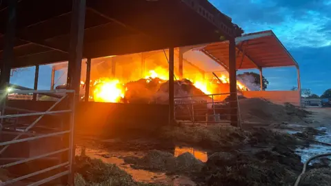 A metal agricultural shed has materials underneath it that are engulfed in bright orange flames 