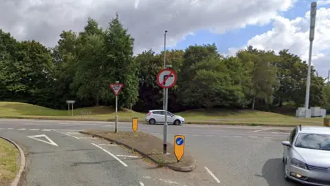 Google Road junction with a no right turn sign and a give way sign from a minor road to a major road. A grey car is turning into the minor road, while a white car is passing on the major road in front of trees.