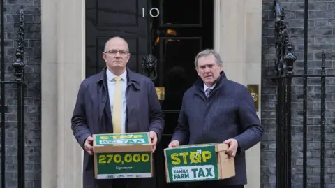 Jonathan Brady/PA Two men in suits and overcoats standing outside the shiny black door of No 10 Downing Street. On the left is NFU President Tom Bradshaw and on the right is NFU Cymru president Aled Jones. Both are holding cardboard boxes that say "stop the family farm tax" and "270,000 signatures" on them. 