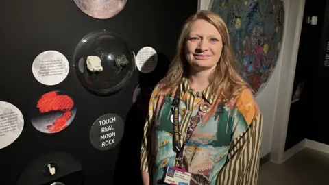 A woman with long light brown hair stands in front of a moon rock exhibition. There are signs on the wall that encourage people to touch part of the exhibition. The woman is wearing a multi-coloured blouse. 