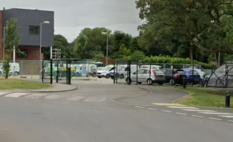 Google A general view of cars in a car park. There is black mesh fencing and there are trees in the background.