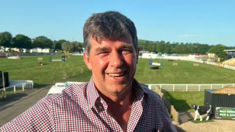 David Cotton smiles at the camera with a showjumping ring behind him.